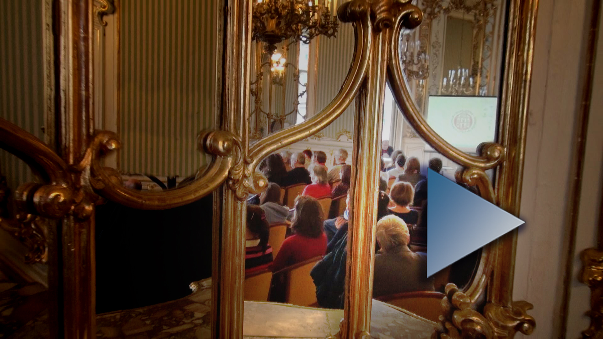 A könyvtár dísztermének rokokó ornamentikájú tükrében a konferencia közönsége látszik. Ha a képre kattint, a videó oldalára jut.